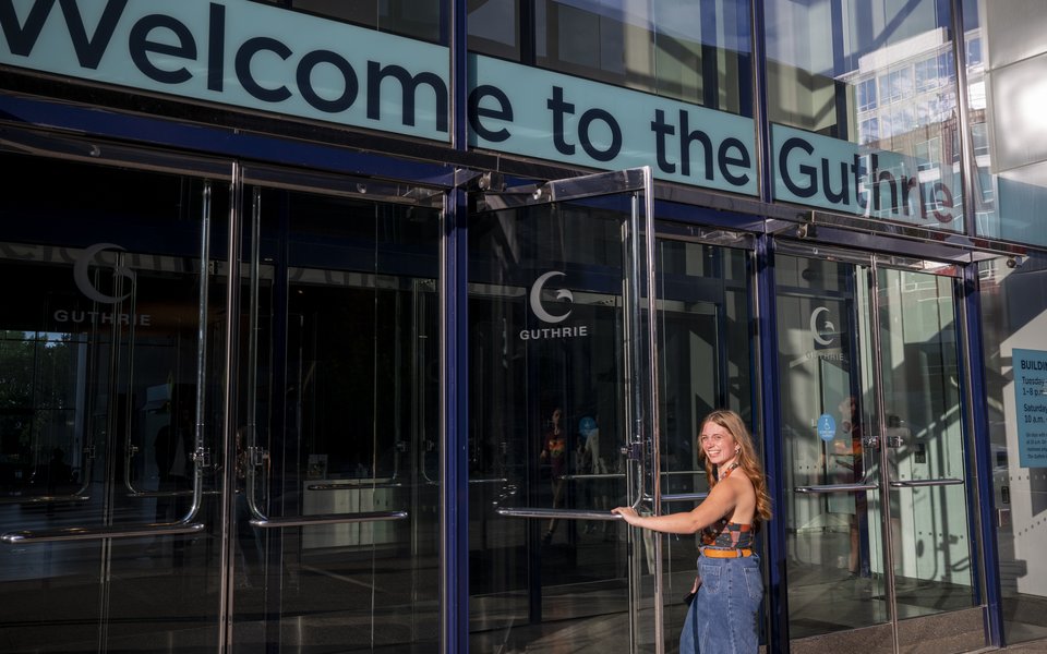 An image of a young woman smiling at the camera as she enters the main doors of the Guthrie Theater building, just under a blue sign that reads "Welcome to the Guthrie"